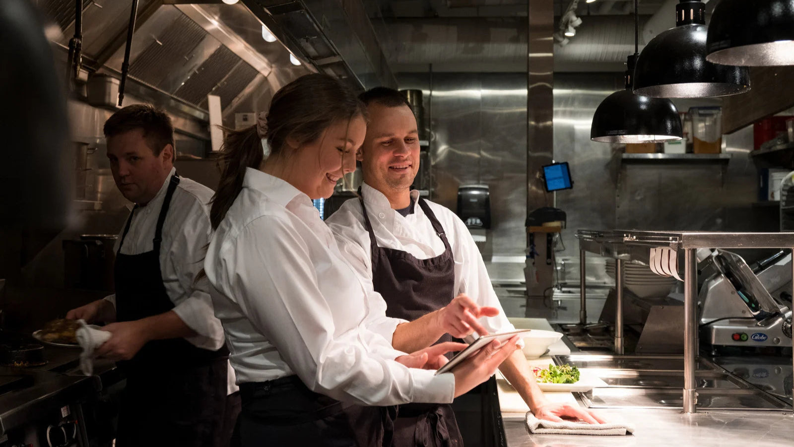 Three chefs in a professional kitchen using a tablet device.